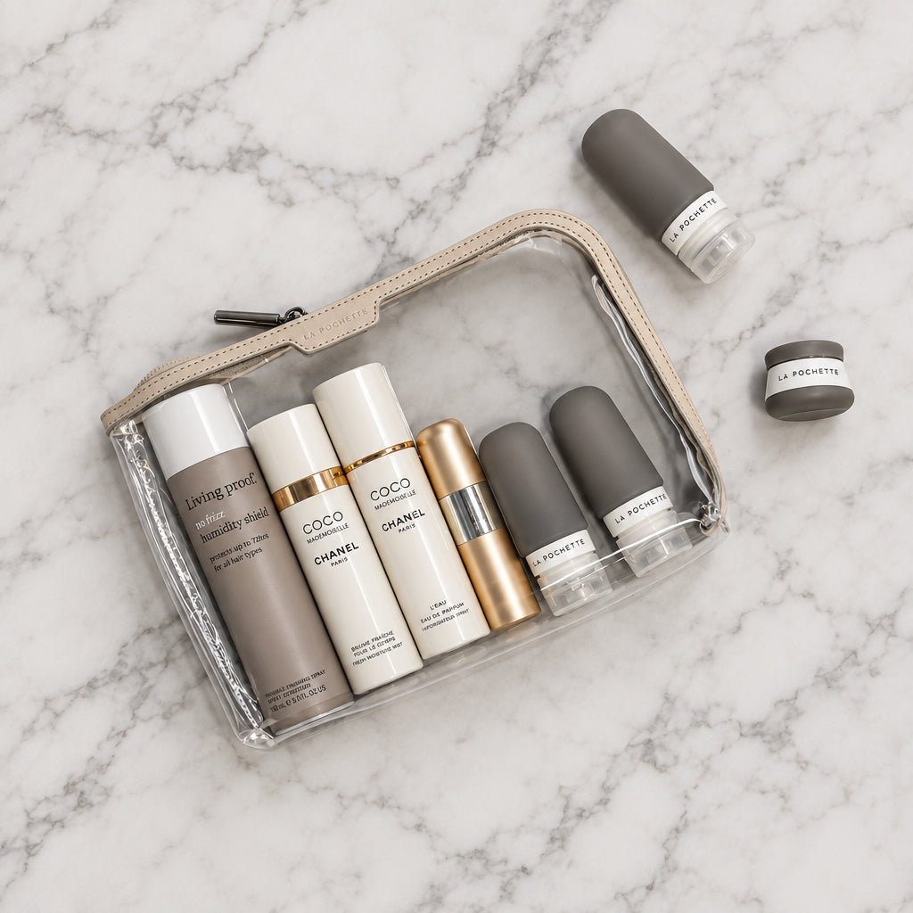 A clear zippered La Pochette pouch with a beige zipper, containing several cosmetic bottles and la Pochette bottles and pots arranged on a marble table top, with a few items placed outside the pouch.