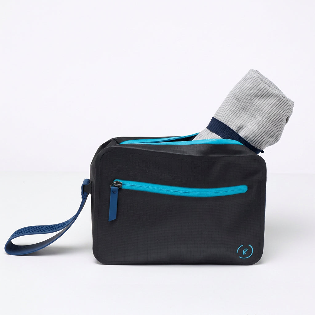 A black la Pochette waterproof Aqua Bag with blue detachable handle and zips with grey microfibre towel rolled up inside bag. On a white background