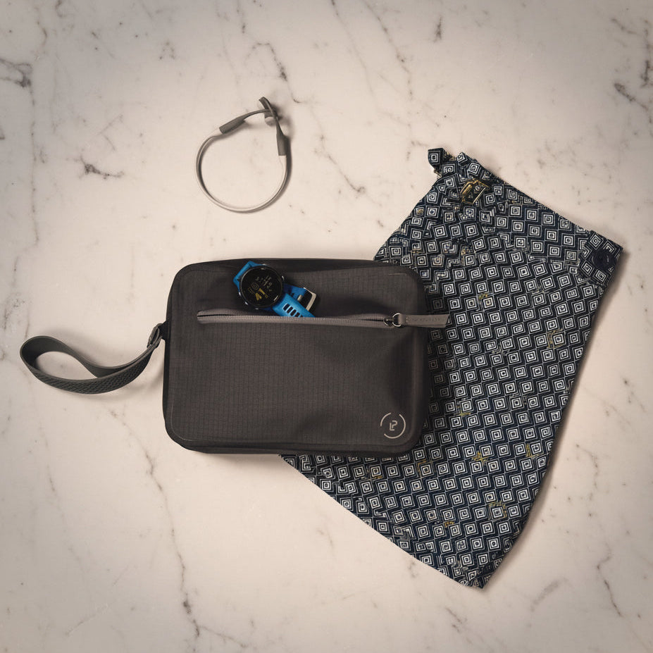 A La Pochette waterproof swim bag with a pair of trunks, sport watch and sports earphones. On a light gray marble surface 
