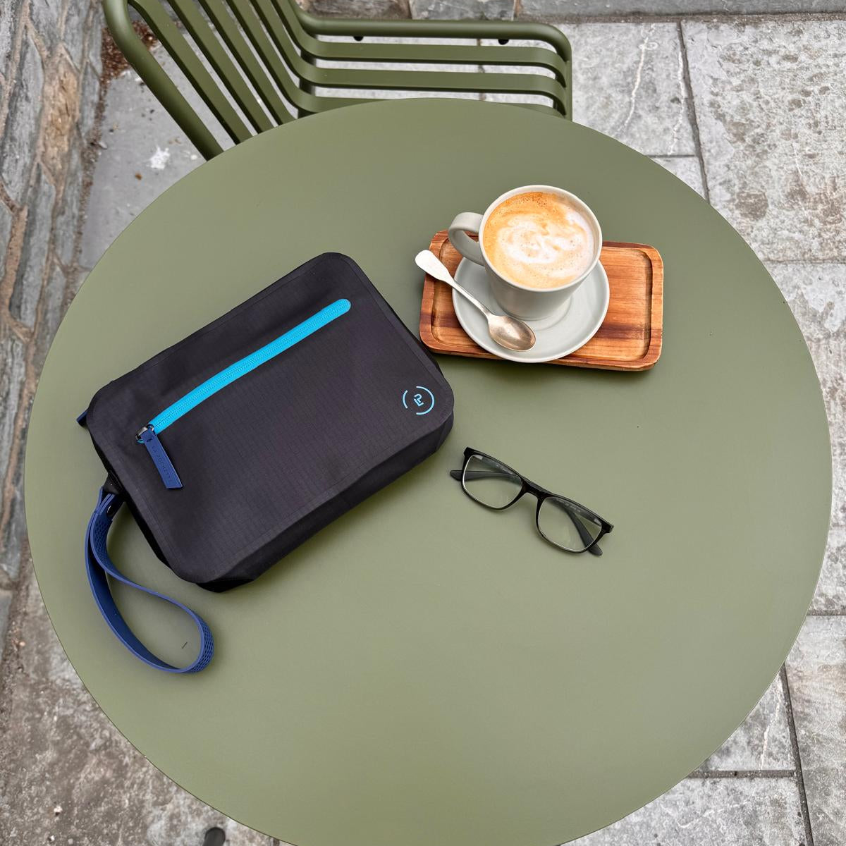 Aqua Bag in black with bright blue trims laying flat on an outside green circular table next to a coffee and a pair of men's glasses
