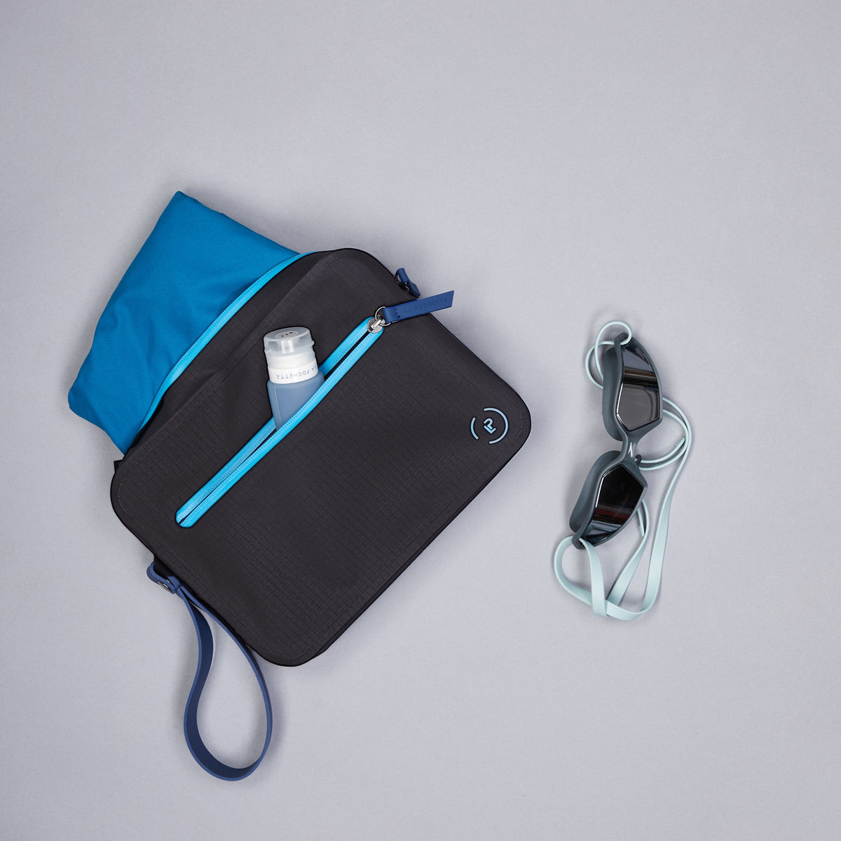 Black waterproof aqua bag with blue details styled with blue swimwear, goggles and a blue La Pochette travel bottle in the separate front pocket