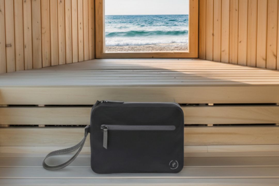 Black waterproof Aqua Bag with dark grey details shown outside a wooden panel sauna overlooking a beach