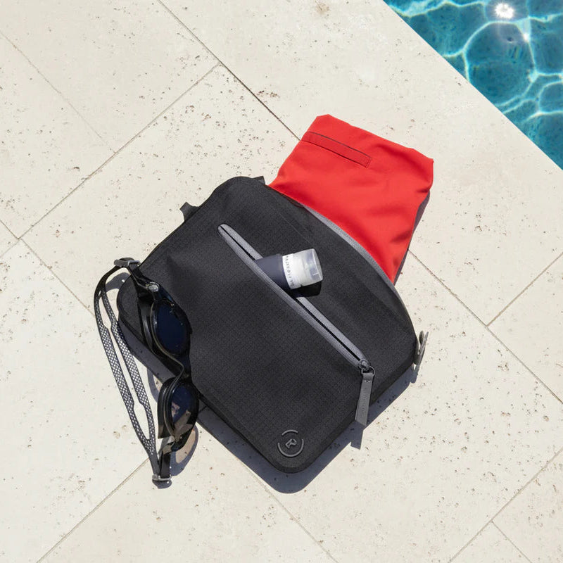 Black waterproof aqua bag with grey details styled with swimwear and goggles on an outside poolside, designed to keep wet and dry items separate