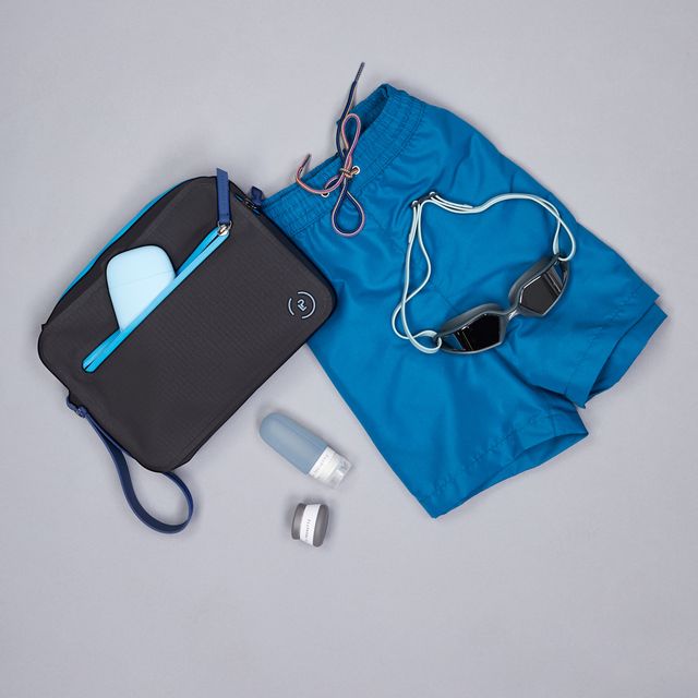 Black waterproof aqua bag with blue details styled with blue swimming shorts, goggles, and the La Pochette travel bottle and pot. Designed to keep wet and dry items separate