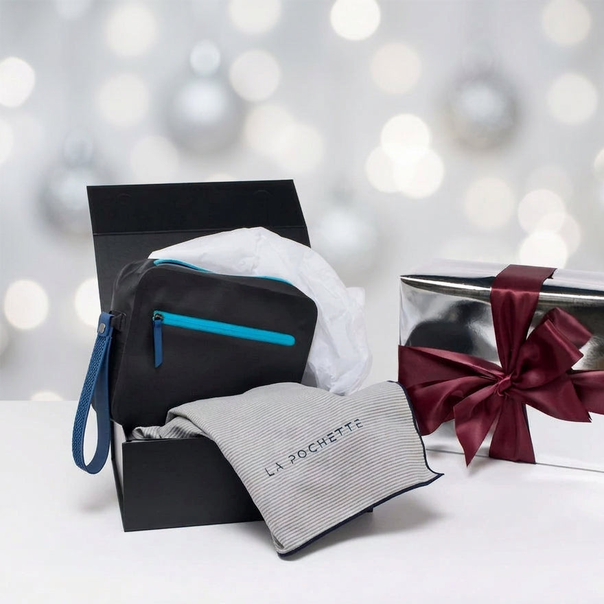 A black La Pochette waterproof Aqua bag with a bright blue zipper and detachable wrist strap, displayed in a black gift box together with a La Pochette branded microfibre grey towel with blue edging. A silver wrapped gift box with a burgundy ribbon bow is next to it on a white background with silver christmas lighting