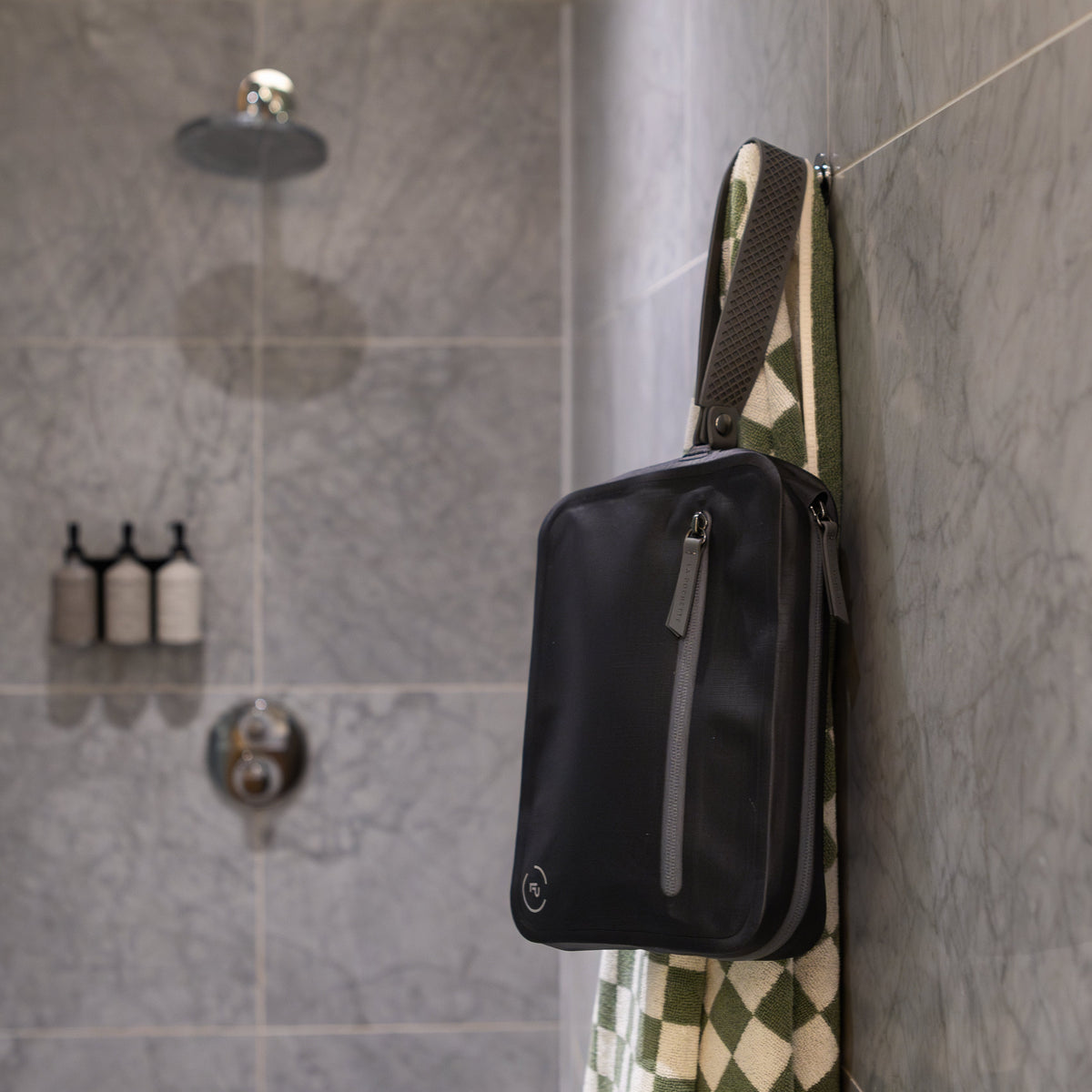 La Pochette Waterproof Aqua Bag in black and grey, hanging on a hook in a shower cubicle with green towel.