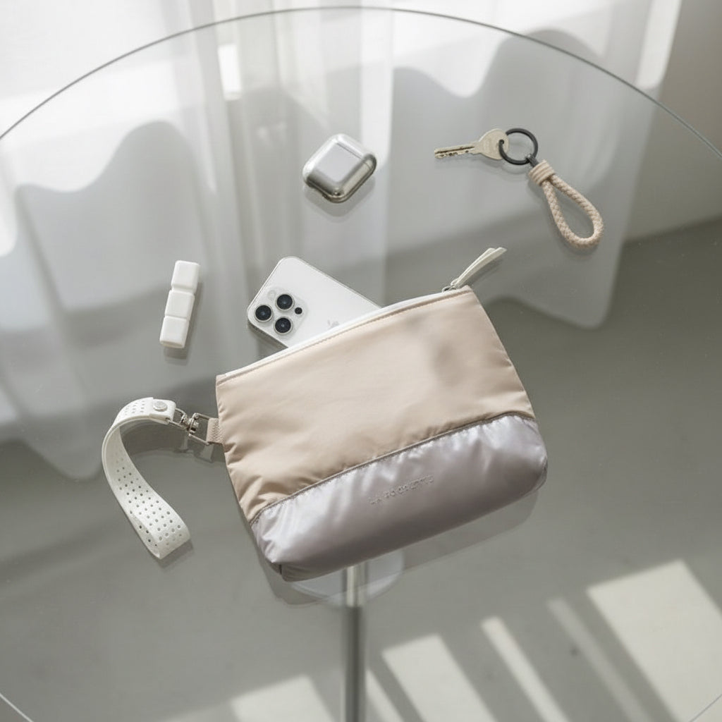 Beige and grey La Pochette clutch bag with pouch with detachable white strap on a glass table with phone, keychain, and other items 