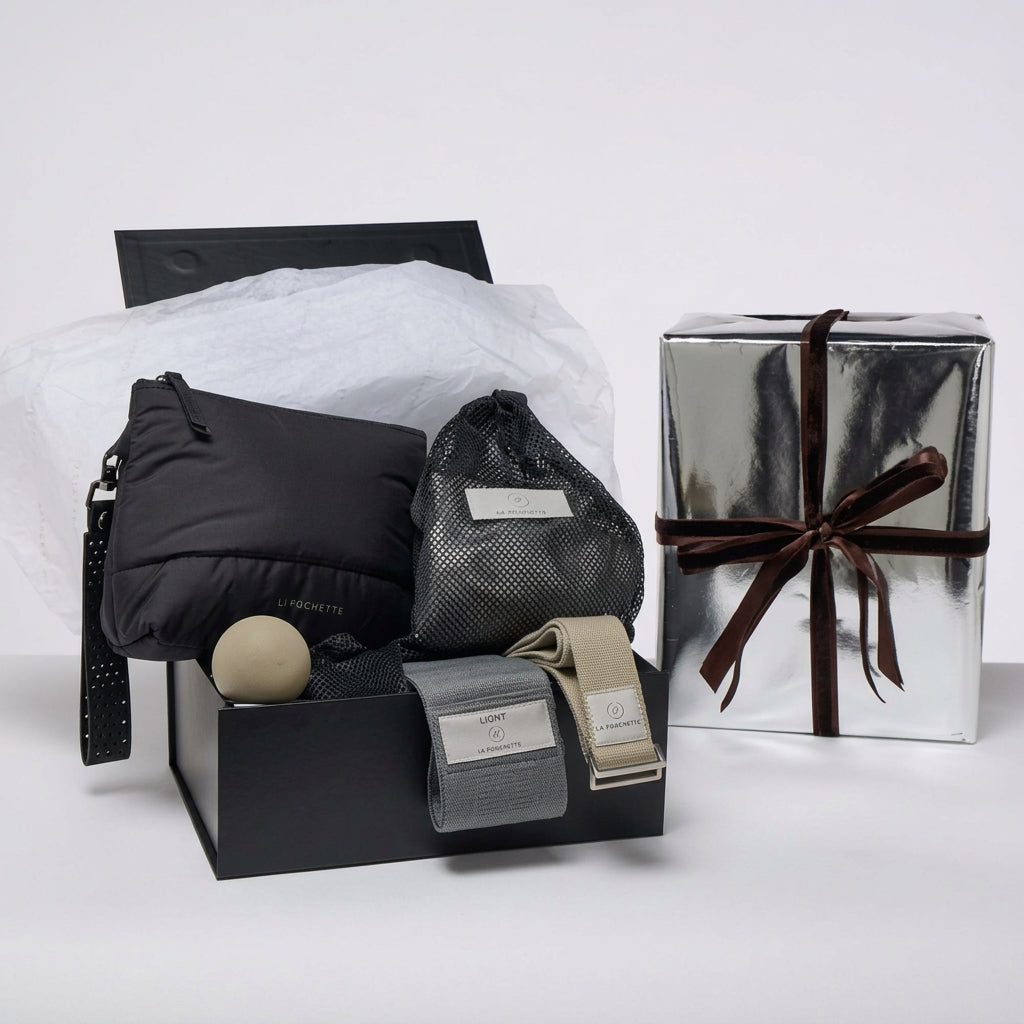 A curated gift set displayed on a white studio background, featuring an open black box with La Pochette workout accessories. Including black and grey pouch with - Detachable handle
- A grey resistance band
- A taupe yoga strap
- A grey small silicone recovery ball
- An ivory inflatable Pilates ball in a black mesh pouch
 a silver gift box with a brown velvet 

