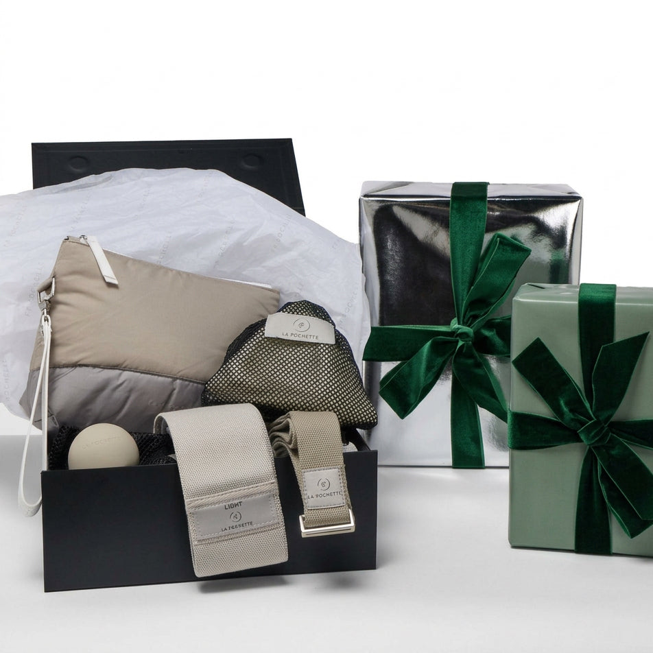 A curated gift set displayed on a white studio background, featuring an open black box with La Pochette workout accessories. Including Taupe and grey pouch with - Detachable handle
- A grey resistance band
- A taupe yoga strap
- A grey small silicone recovery ball
- An ivory inflatable Pilates ball in a black mesh pouch
 a silver gift box with a dark green ribbon, and a sage green gift box with a matching ribbon are at the side 
