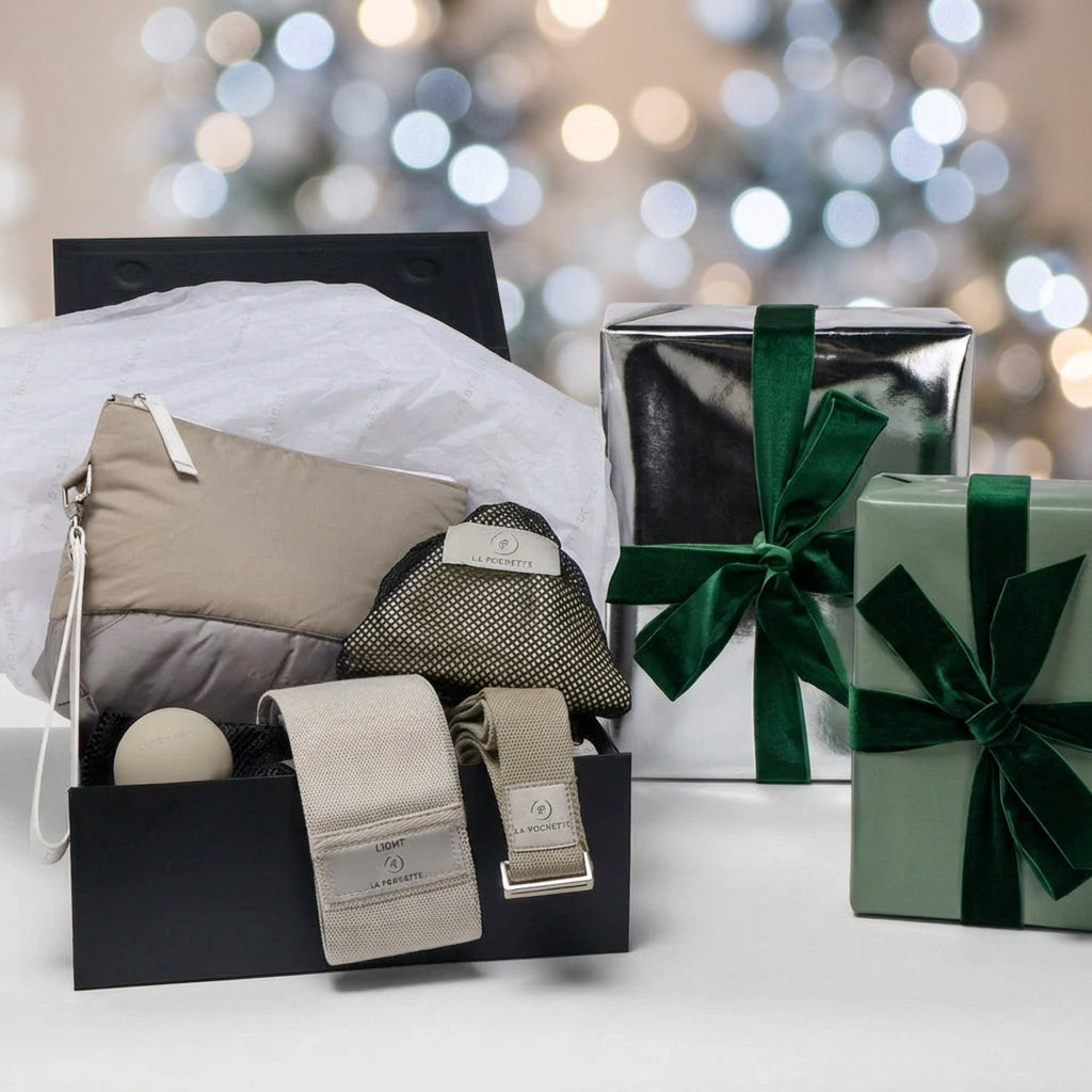 A curated gift set displayed, featuring an open black box with La Pochette workout accessories. Including Taupe and grey pouch with - Detachable handle
- A grey resistance band
- A taupe yoga strap
- A grey small silicone recovery ball
- An ivory inflatable Pilates ball in a black mesh pouch
 a silver gift box with a dark green ribbon, and a sage green gift box with a matching ribbon are at the side 

Blurred Christmas lights in the background 
