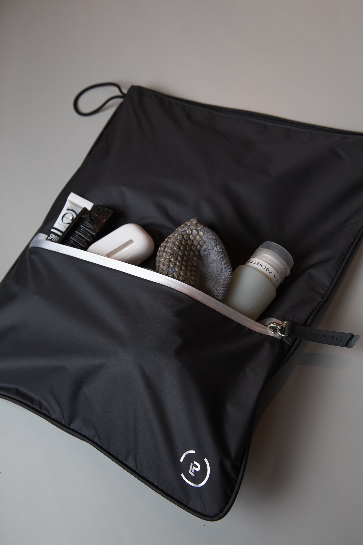 Black La Pochette Sweat Bag with silver zip with items including la pochette silicone travel bottle, grip socks, AirPods in the front pocket