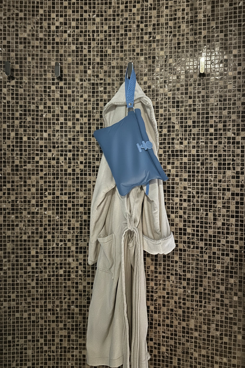 A beige towelling robe  hung with a blue Large La Pochette Waterproof bag in a spa . dark mosaic tile wall background