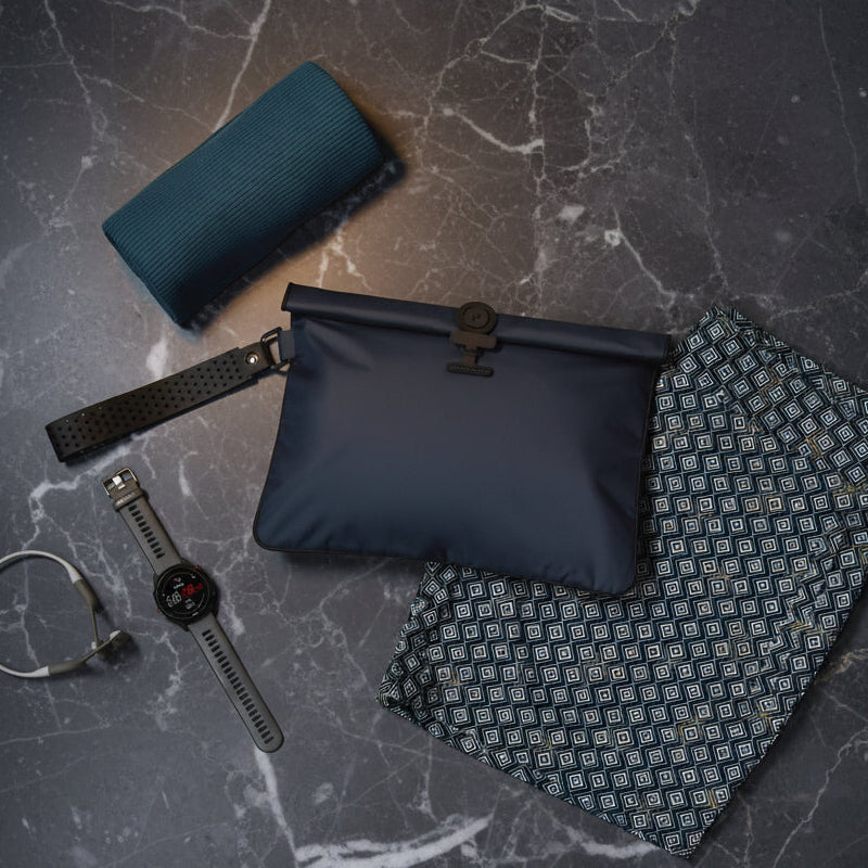 A flat-lay product shot of a navy blue La Pochette waterproof wet bag with a silicone black detachable handle and  clasp.  Shown with its blue patterned trunks, rolled up micro fibre towel, a gray smartwatch on a  grey marble background
