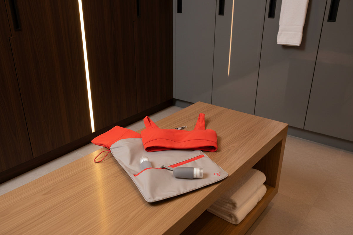 Taupe and red sweat bag, shown on a bench in  a gym changing room, with red gym kit and la pochette travel bottles