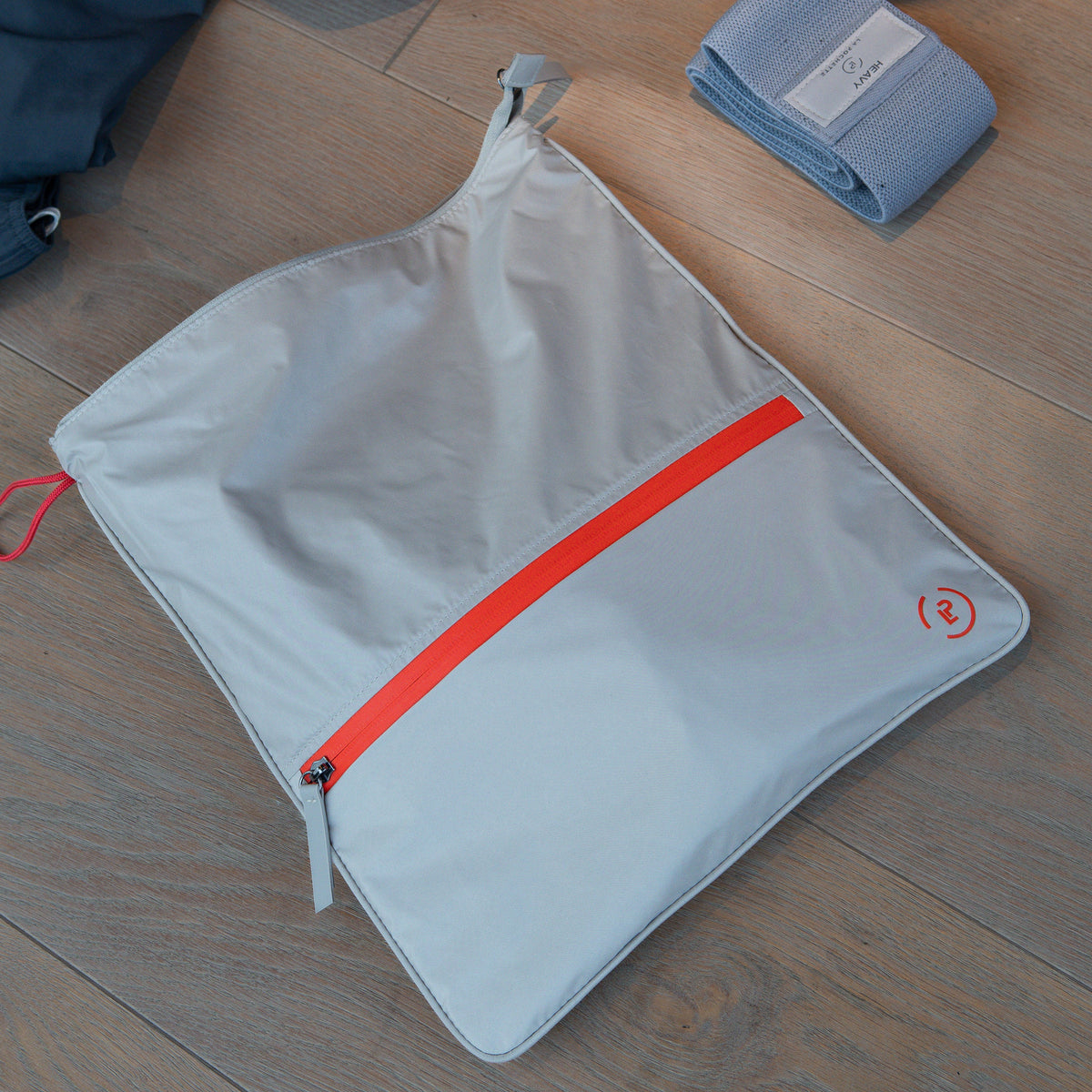 Beige La Pochette Antimicrobial Gym Bag with red trims next to a La Pochette resistance band on a wooden floor