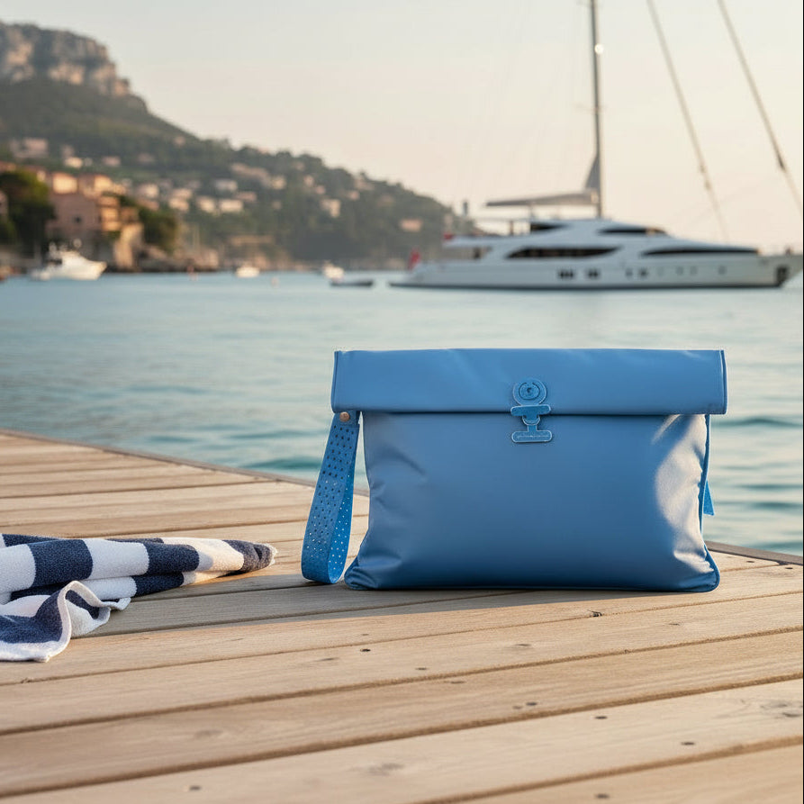A la Pochette Waterproof wetbag in blue on a deck with a yacht in the background. Also shown is a striped blue and white towel. 