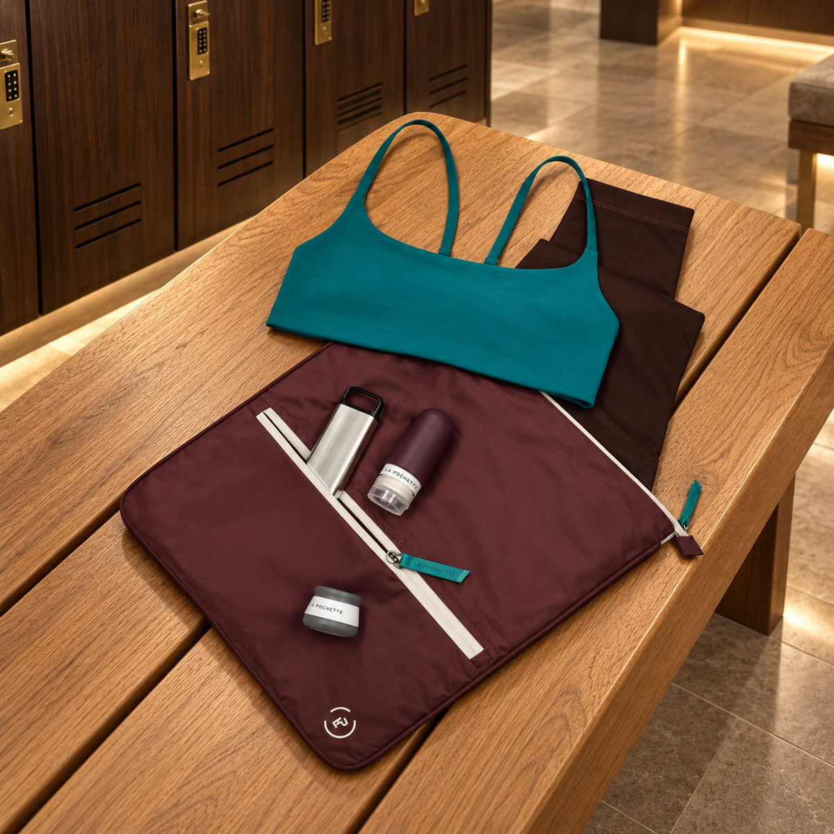 A teal sports bra and dark brown leggings are laid out on a wooden bench next to a  burgundy and teal La Pochette Sweat bag  holding La Pochette travel bottles and pots with locker doors in the background.