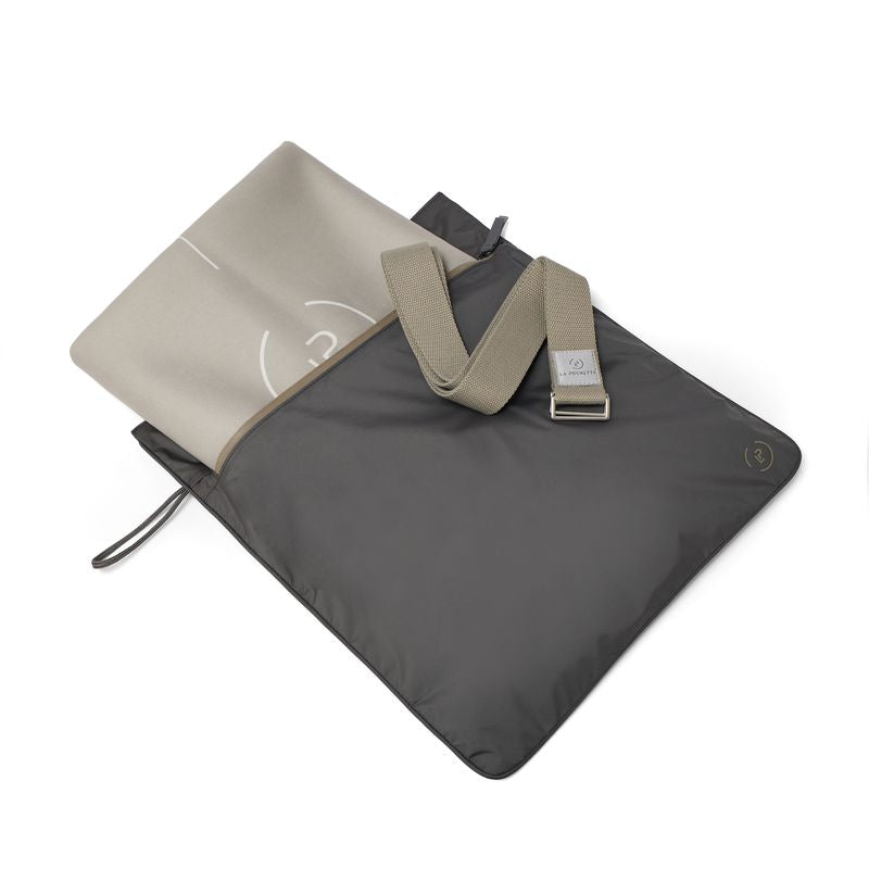 A dark waterproof La Pochette Kit Bag with a beige and white lightweight travel yoga mat  inside, and a Taupe brown La Pochette yoga strap laid on top. All on  a white surface.