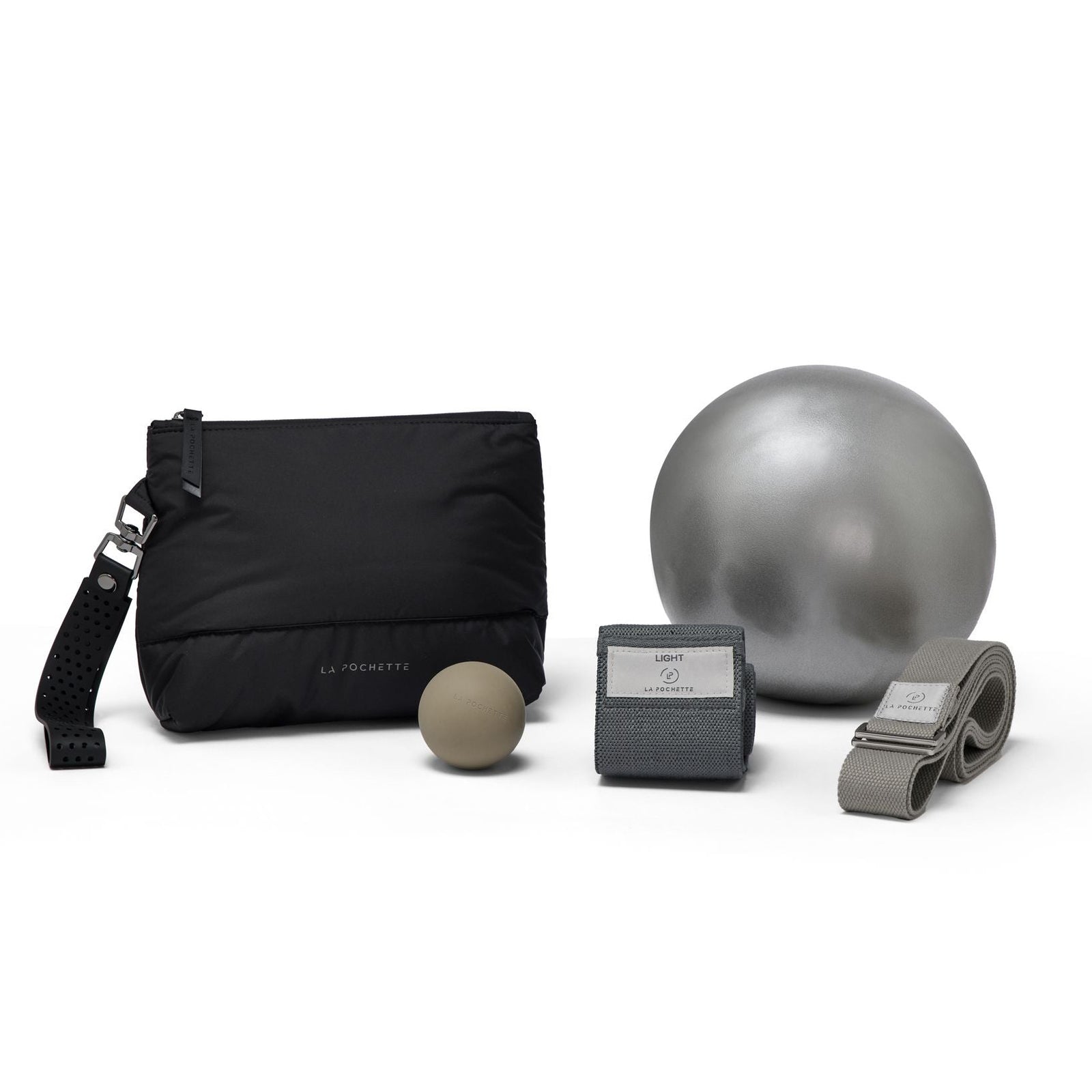 An assortment of workout accessories in black and grey, including:
- an inflatable Pilates ball in silver
- a grey yoga strap with a woven logo saying La Pochette
- a resistance band with a woven logo saying La Pochette
- a grey small silicone recovery ball
- a La Pochette Everywhere pouch in black with black silicone handle

On a white background 