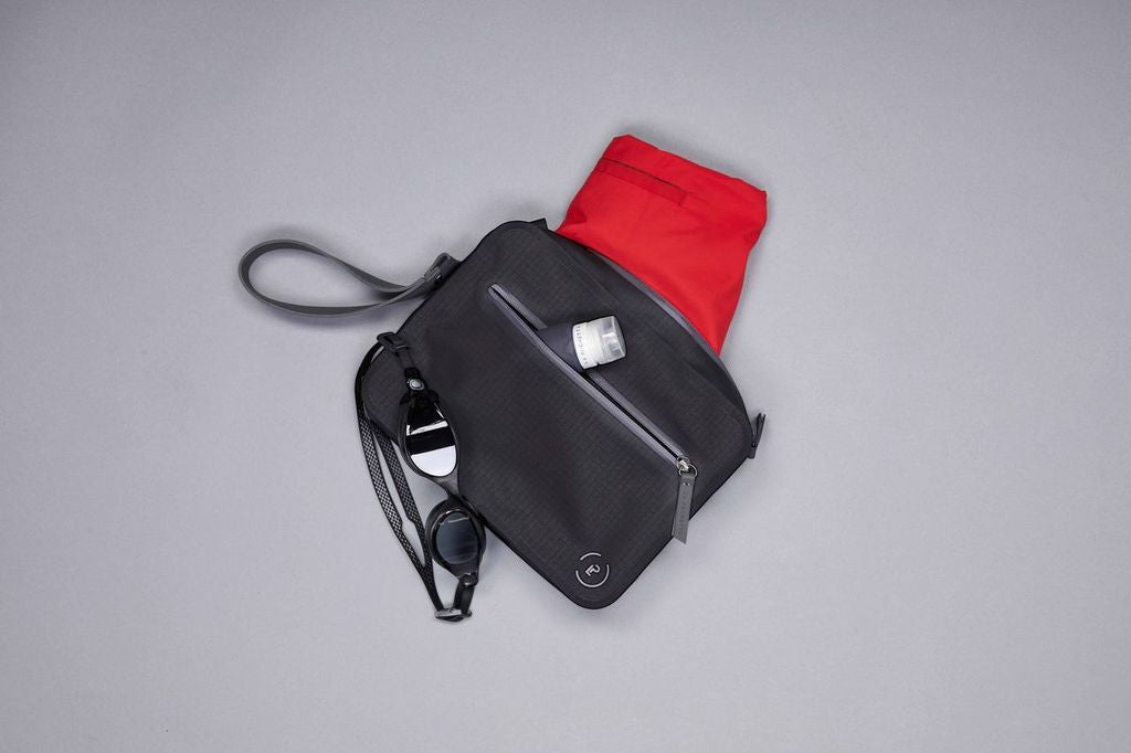 Aqua Bag in Black and Pewter, shown with red trunks in main pocket, and La Pochette travel bottle in front pocket.