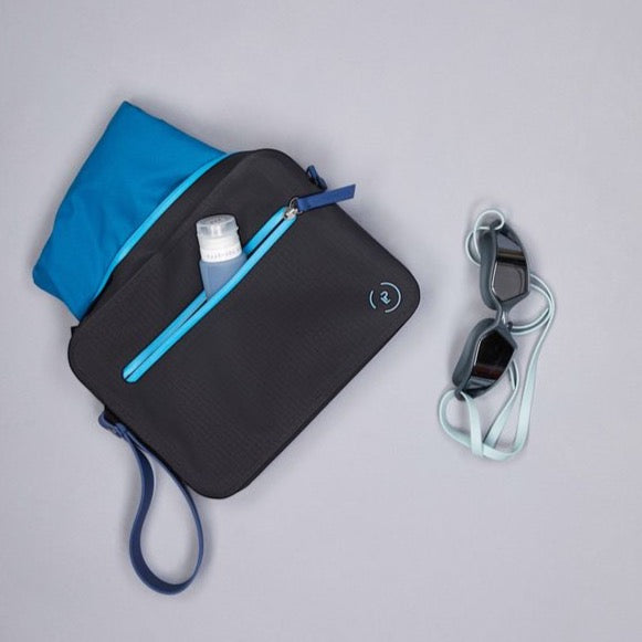 Aqua Bag in black and marine blue. Shown with trunks inside main pocket, goggles on the side, and La Pochette travel bottles in front pocket