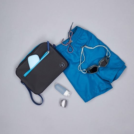 Aqua Bag in black and marine blue. Shown with trunks, goggles, and La Pochette travel bottles and pots.