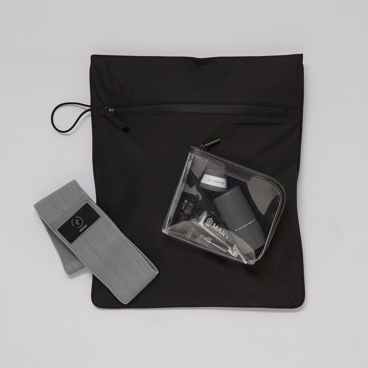Black Kit Bag laying flat with a La Pochette clear cosmetic pouch holding essentials and a grey La Pochette Resistance Band laying over it 
