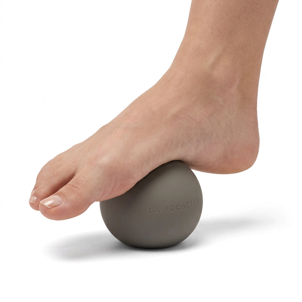 A matte taupe brown silicone ball with the brand name 'LA POCHETTE' embossed on the front, underneath a woman's foot. Being rolled on the arches. Against a white background 