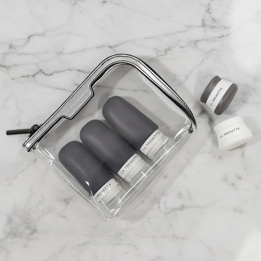 Small clear travel bag in silver and black with refillable La Pochette pots and bottles on a marble table
