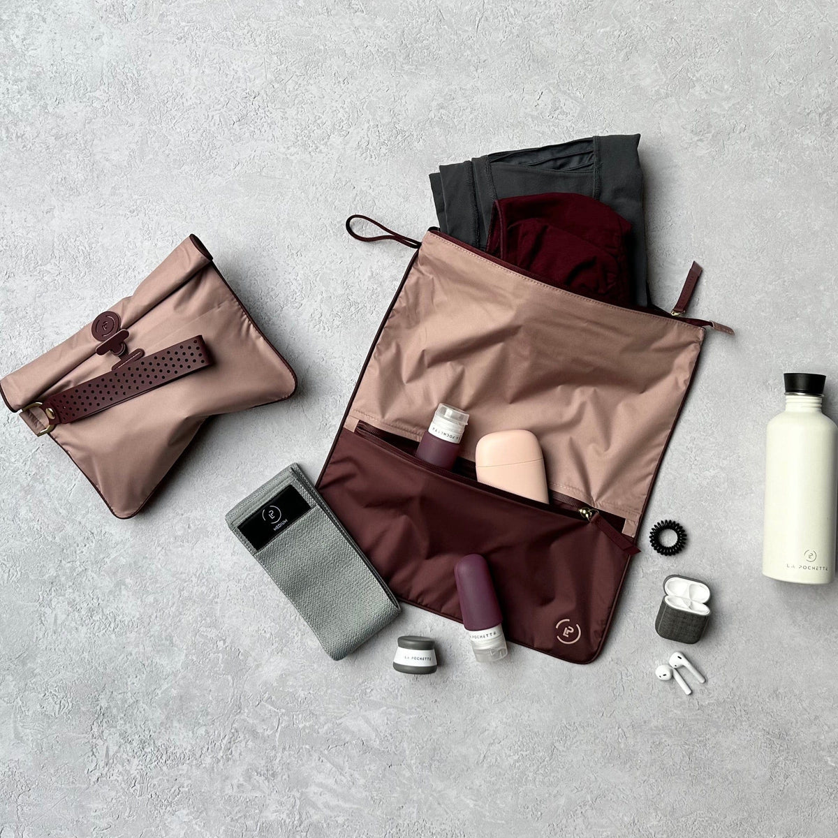 Soft pink and burgundy Sweat Bag shown flat styled with gym essentials including the La Pochette travel bottle, resistance band, water bottle, and Small Wet Bag