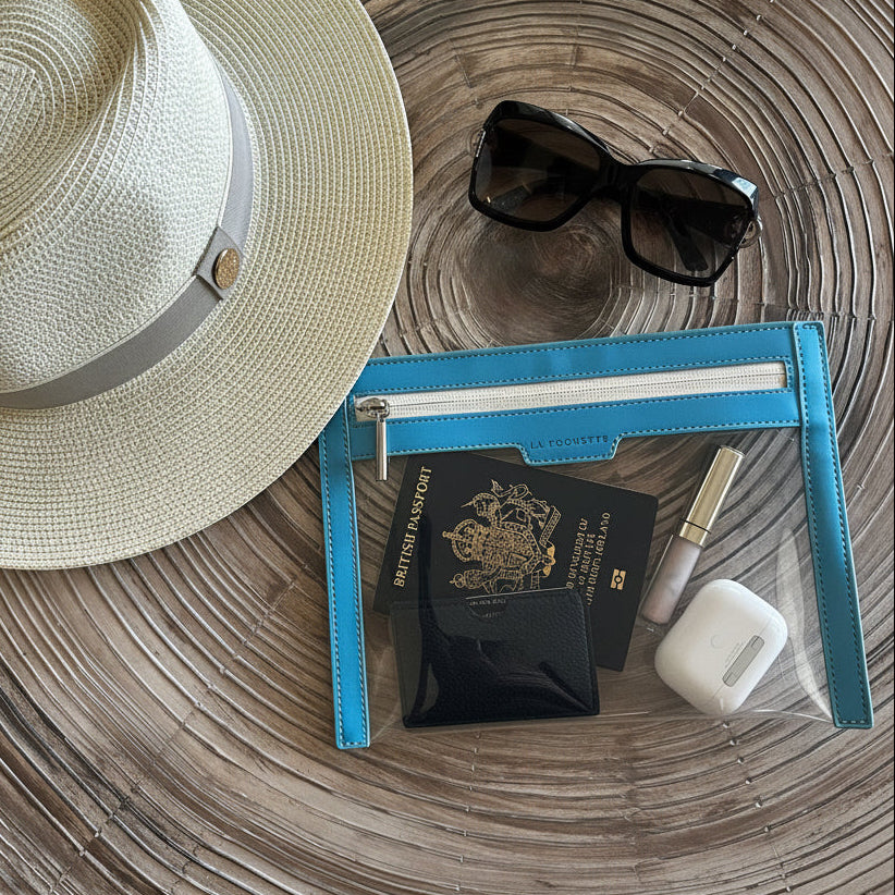 A styled flat-lay of a straw fedora hat with a gray band, black sunglasses, a bright blue transparent La Pochette wallet with white zip. Inside wallet is a UK passport, a lip balm, and a white EarPods case. All  on a round wooden table.