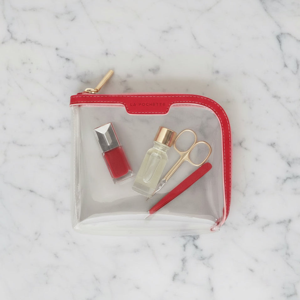 Small clear cabin approved La Pochette Pouch with red and pink trim, containing red nail polish, nail scissors, and red tweezers.