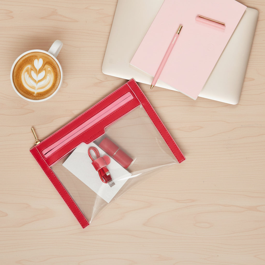 Clear La Pochette wallet with red trims and pink zip on a desk next to laptop and coffee. Red lipstick case, and red la pochette cable tidy in pouch.