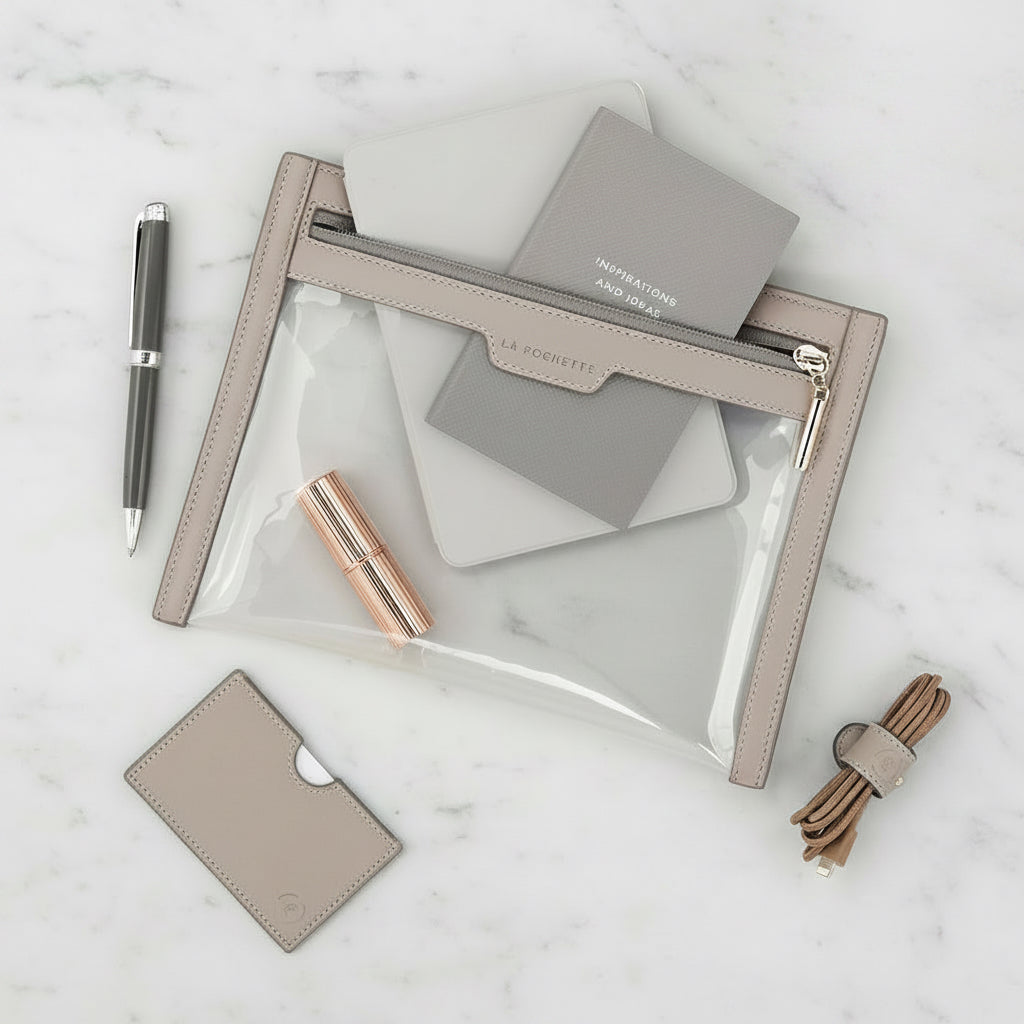 La Pochette clear wallet,  in taupe colourway Notebook, lipstick, and kindle inside the wallet. On a marble table next to la Pochette cable tie and card wallet. 