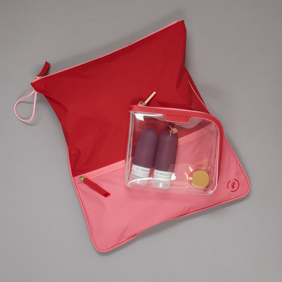 Vibrant pink and red Sweat Bag laid flat, topped with a small La Pochette clear cosmetic Bag holding La Pochette silicone travel bottles.